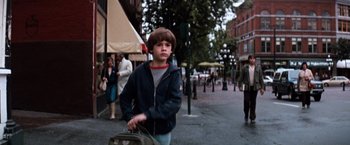 Movie still from “The NeverEnding Story” (1984), directed by Wolfgang Petersen – A boy is walking down the street holding a bag; Medium shot, Over the shoulder angle