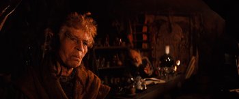 Movie still from “The NeverEnding Story” (1984), directed by Wolfgang Petersen – A man with a long hair and a red beard is sitting in front of a counter; Close Up shot, Over the shoulder angle