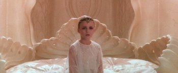 Movie still from “The NeverEnding Story” (1984), directed by Wolfgang Petersen – A young girl in a white dress sitting in front of a bed; Medium shot, High angle