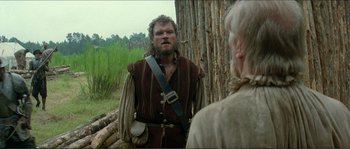 Movie still from “The New World” (2005), directed by Terrence Malick – A man in a brown and black outfit standing next to a tree; Medium shot, Over the shoulder angle