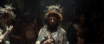 Movie still from “The New World” (2005), directed by Terrence Malick – A man with a feather headdress is standing in front of a group of people; Medium shot, Over the shoulder angle