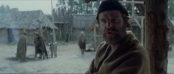 Movie still from “The New World” (2005), directed by Terrence Malick – A man with a long beard and a black hat is standing in front of a wooden building; Medium shot, Over the shoulder angle