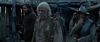 Movie still from “The New World” (2005), directed by Terrence Malick – An old man with long hair and a beard is standing in front of a group of people; Medium shot, Over the shoulder angle