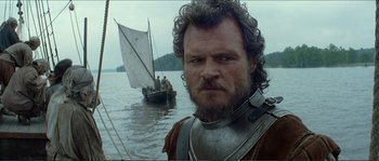 Movie still from “The New World” (2005), directed by Terrence Malick – A man with a beard and a helmet on standing in front of a sail boat; Close Up shot, Low angle