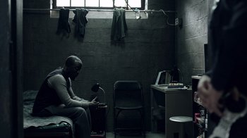 Movie still from “The Night Of” (2016), directed by James Marsh – A man sitting in a room next to a desk; Medium shot, High angle