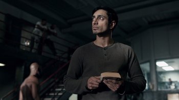Movie still from “The Night Of” (2016), directed by James Marsh – A man holding a book while standing in front of a staircase; Medium shot, Low angle