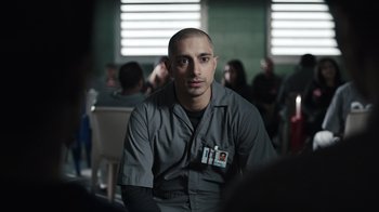 Movie still from “The Night Of” (2016), directed by James Marsh – A man with a badge around his neck is sitting in front of a group of people; Close Up shot, Over the shoulder angle
