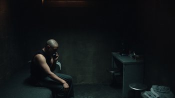 Movie still from “The Night Of” (2016), directed by James Marsh – A man sitting on a couch talking on a phone; Wide shot, High angle