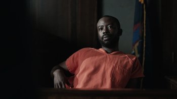 Movie still from “The Night Of” (2016), directed by James Marsh – A man in an orange shirt sitting in front of a wooden table; Medium shot, Low angle