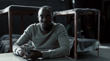 Movie still from “The Night Of” (2016), directed by James Marsh – A man sitting at a table in front of a bunk bed; Medium shot, Over the shoulder angle