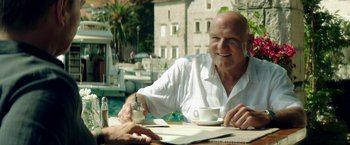 Movie still from “The November Man” (2014), directed by Roger Donaldson – An older man sitting at a table with a cup of coffee; Medium shot, Over the shoulder angle