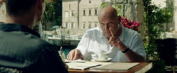 Movie still from “The November Man” (2014), directed by Roger Donaldson – An older man sitting at a table with a cup of coffee; Medium shot, Over the shoulder angle
