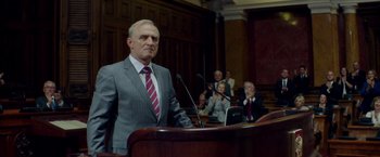 Movie still from “The November Man” (2014), directed by Roger Donaldson – A man standing at a podium in front of microphones; Medium shot, Low angle