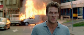Movie still from “The November Man” (2014), directed by Roger Donaldson – A man standing in front of a burning car; Close Up shot, Over the shoulder angle