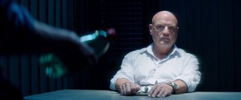 Movie still from “The November Man” (2014), directed by Roger Donaldson – A man sitting at a table holding a pair of scissors in front of a bottle; Medium shot, Low angle