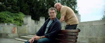 Movie still from “The November Man” (2014), directed by Roger Donaldson – A man sitting on top of a wooden park bench next to another man; Medium shot, Over the shoulder angle