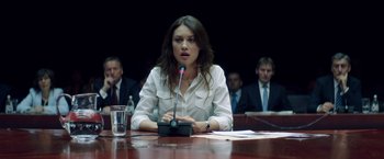 Movie still from “The November Man” (2014), directed by Roger Donaldson – A woman sitting in front of a microphone at a table; Medium shot, Over the shoulder angle
