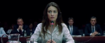 Movie still from “The November Man” (2014), directed by Roger Donaldson – A woman is holding a microphone in front of a crowd; Close Up shot, Over the shoulder angle