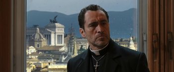 Movie still from “The Nun” (2018), directed by Corin Hardy – A man wearing a priest's outfit and a cross necklace; Close Up shot, Low angle