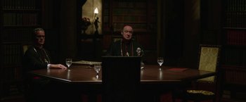 Movie still from “The Nun” (2018), directed by Corin Hardy – A priest is sitting at a table in front of a microphone; Medium shot, Over the shoulder angle