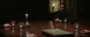 Movie still from “The Nun” (2018), directed by Corin Hardy – Two men sitting at a table in a dark room; Medium shot, Over the shoulder angle