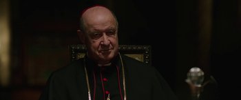 Movie still from “The Nun” (2018), directed by Corin Hardy – An older man wearing a red and black outfit; Close Up shot, Over the shoulder angle