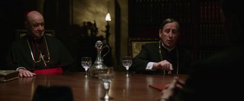 Movie still from “The Nun” (2018), directed by Corin Hardy – A man sitting at a wooden table in front of a wine decanter; Medium shot, Over the shoulder angle