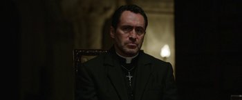 Movie still from “The Nun” (2018), directed by Corin Hardy – A man wearing a priest's outfit sitting in a chair; Close Up shot, Low angle