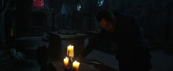 Movie still from “The Nun” (2018), directed by Corin Hardy – A man standing in front of a table with lit candles on top of it; Medium shot, Low angle