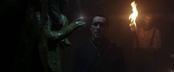 Movie still from “The Nun” (2018), directed by Corin Hardy – A man standing in front of a statue of a creature; Medium shot, Low angle