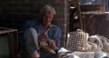 Movie still from “The Nutty Professor” (1996), directed by Tom Shadyac – An older man sitting on the ground smoking a cigarette; Medium shot, High angle