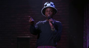 Movie still from “The Nutty Professor” (1996), directed by Tom Shadyac – A man wearing a hat and holding a microphone in front of a brick wall; Medium shot, Low angle
