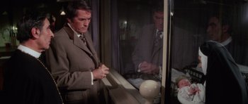 Movie still from “The Omen” (1976), directed by Richard Donner – A man standing in front of a window looking at himself in the mirror; Medium shot, High angle