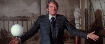 Movie still from “The Omen” (1976), directed by Richard Donner – A man wearing a suit and tie standing in front of a painting; Medium shot, Low angle
