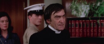 Movie still from “The Omen” (1976), directed by Richard Donner – A man wearing a priest's outfit and a white hat; Close Up shot, Over the shoulder angle