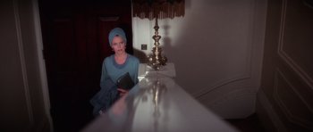 Movie still from “The Omen” (1976), directed by Richard Donner – A woman sitting on the edge of a counter holding a book; Medium shot, Over the shoulder angle