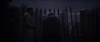 Movie still from “The Omen” (1976), directed by Richard Donner – Two people standing next to each other in front of a metal fence; Medium shot, Low angle