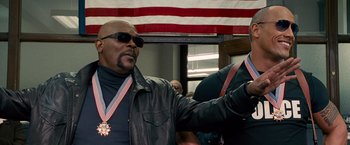 Movie still from “The Other Guys” (2010), directed by Adam McKay – A man in a leather jacket and sunglasses holding a medal in front of an american flag; Medium shot, Low angle