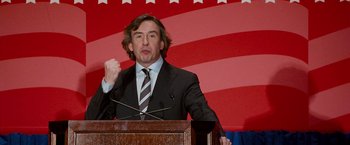 Movie still from “The Other Guys” (2010), directed by Adam McKay – A man in a suit and tie speaking at a podium; Medium shot, Low angle