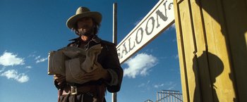 Movie still from “The Outlaw Josey Wales” (1976), directed by Clint Eastwood – A man in a cowboy hat holding a bag under a street sign; Medium shot, Low angle