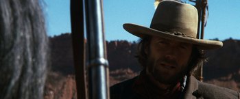 Movie still from “The Outlaw Josey Wales” (1976), directed by Clint Eastwood – A man with a cowboy hat and a rifle in the background; Close Up shot, Low angle