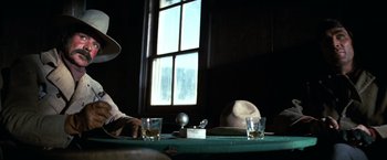 Movie still from “The Outlaw Josey Wales” (1976), directed by Clint Eastwood – A man sitting at a table with a hat on it; Medium shot, Low angle