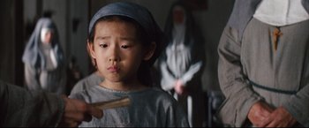 Movie still from “The Painted Veil” (2006), directed by John Curran – A young boy in a nun's outfit holding a wooden stick; Close Up shot, Over the shoulder angle