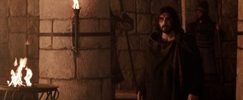 Movie still from “The Passion of the Christ” (2004), directed by Mel Gibson – A man with a beard and a long beard standing in front of a brick wall; Medium shot, Low angle