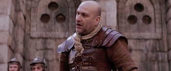 Movie still from “The Passion of the Christ” (2004), directed by Mel Gibson – A bald man in a leather outfit is standing in front of a stone wall; Medium shot, Low angle