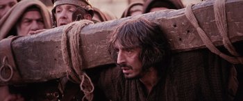 Movie still from “The Passion of the Christ” (2004), directed by Mel Gibson – A man with long hair and beard carrying a large log on his back; Close Up shot, Over the shoulder angle
