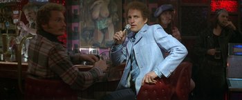 Movie still from “The People vs. Larry Flynt” (1996), directed by Milos Forman – A man sitting in front of a microphone in front of a painting; Medium shot, Low angle