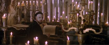 Movie still from “The Phantom of the Opera” (2004), directed by Joel Schumacher – A man wearing a white mask holding a sheet of paper in front of a group of lit candles; Wide shot, High angle