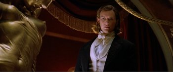 Movie still from “The Phantom of the Opera” (2004), directed by Joel Schumacher – A man in a suit and tie standing next to a table; Close Up shot, Low angle