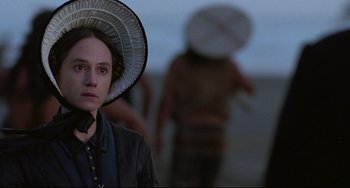 Movie still from “The Piano” (1993), directed by Jane Campion – A woman with a hat is standing in front of a group of people; Close Up shot, Over the shoulder angle